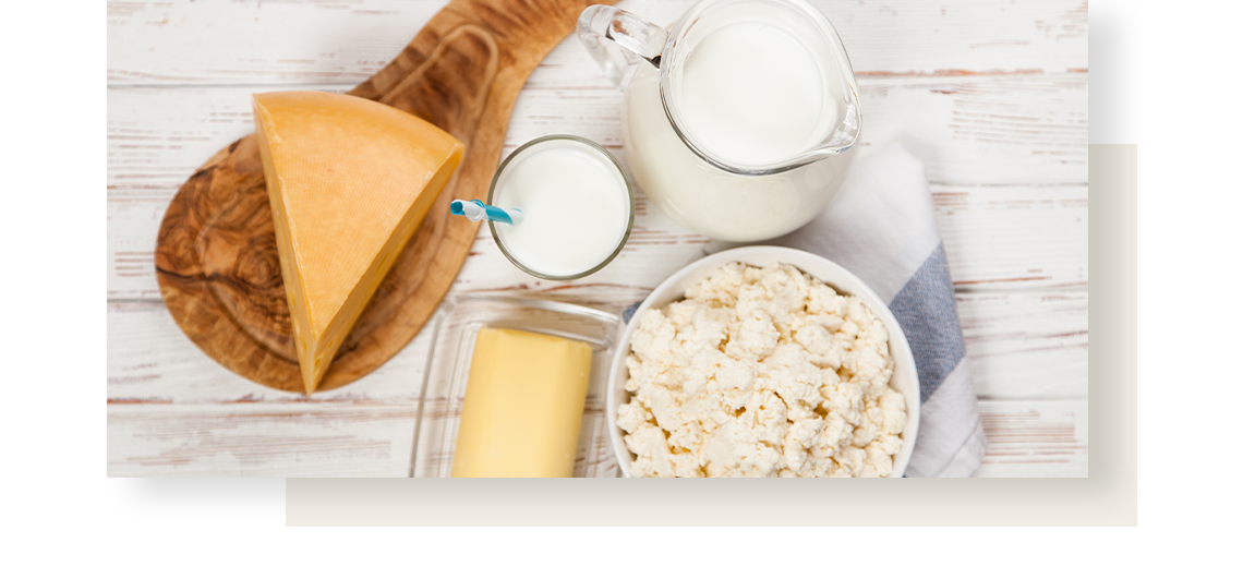 Млечни продукти върху бяла дървена маса: сирене върху дъска, масло, мляко в чаша и кана, извара в купа.